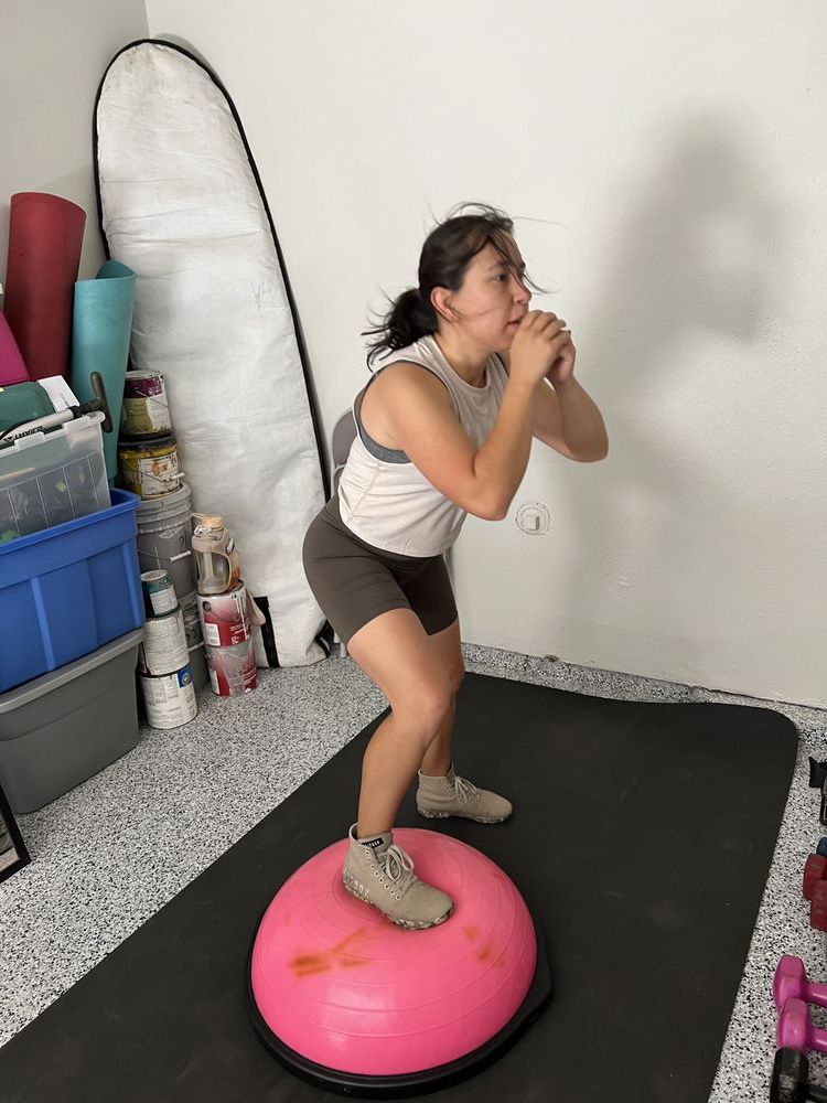 A woman is squatting on a pink exercise ball in a garage.
