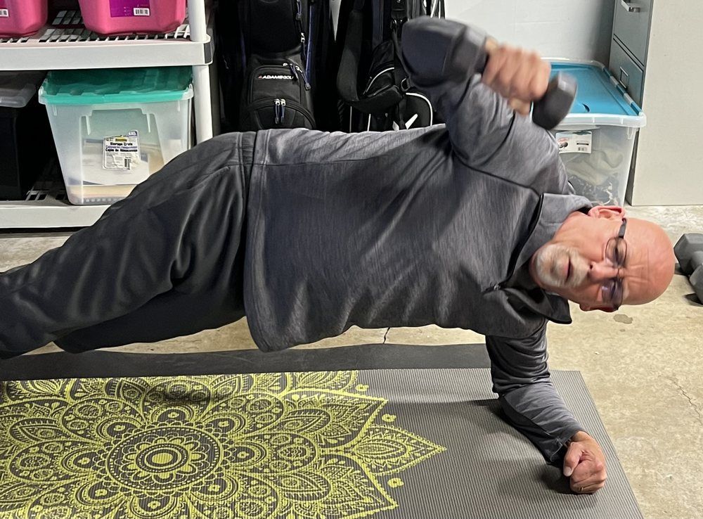A man is doing a plank on a yoga mat while holding a dumbbell.