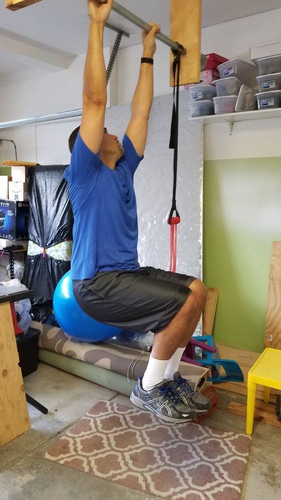 A man is squatting on a blue exercise ball while doing a pull up.