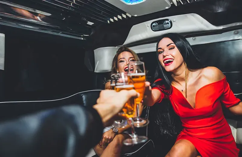 Two women and a person in a limo cheersing beer; woman in red dress smiles and laughs.