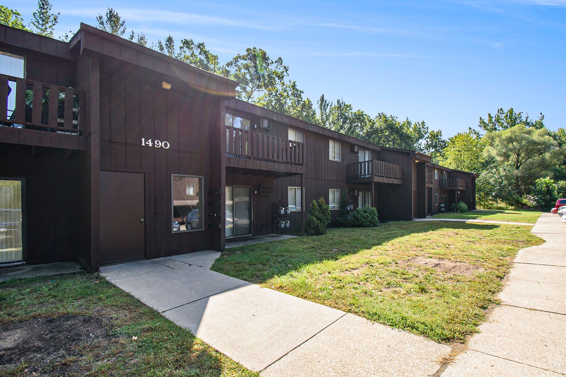Brown two-story apartment building with balconies, concrete sidewalk, and grassy lawn under a blue sky.