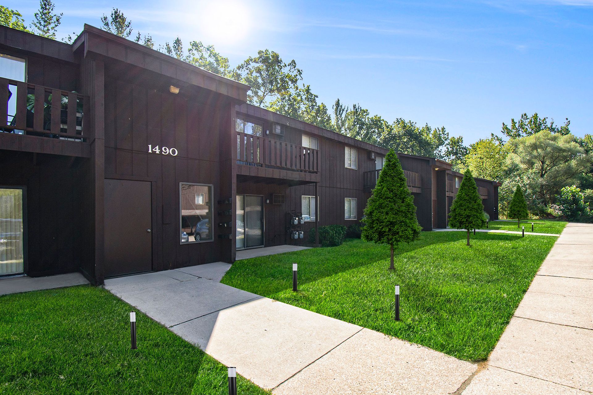 Brown two-story apartment building with balconies, green lawn, and a sunny sky.