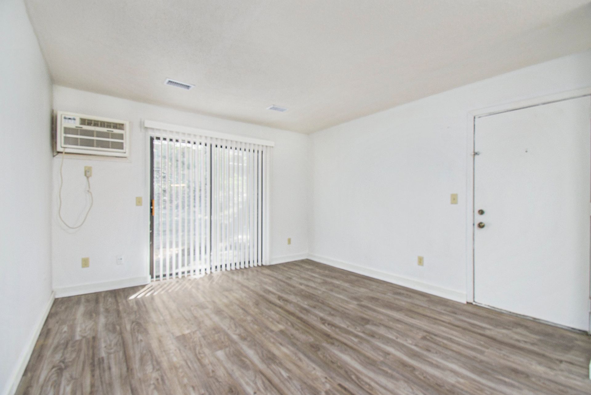 Empty, white-walled room with gray wood-look flooring, sliding glass door with blinds, and air conditioning unit.