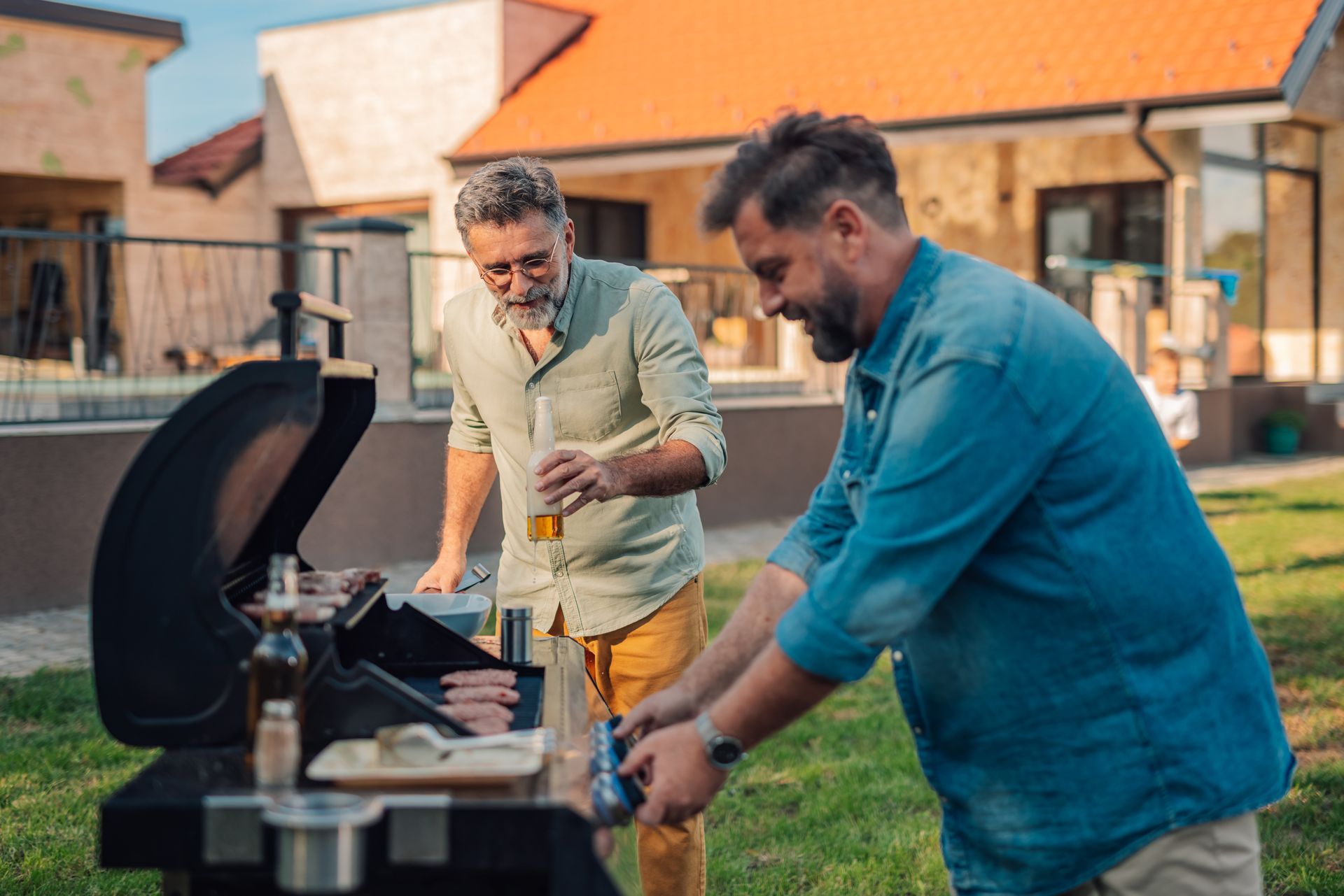 Two men grilling outdoors in a backyard; one pours a beer, one flips food on grill.