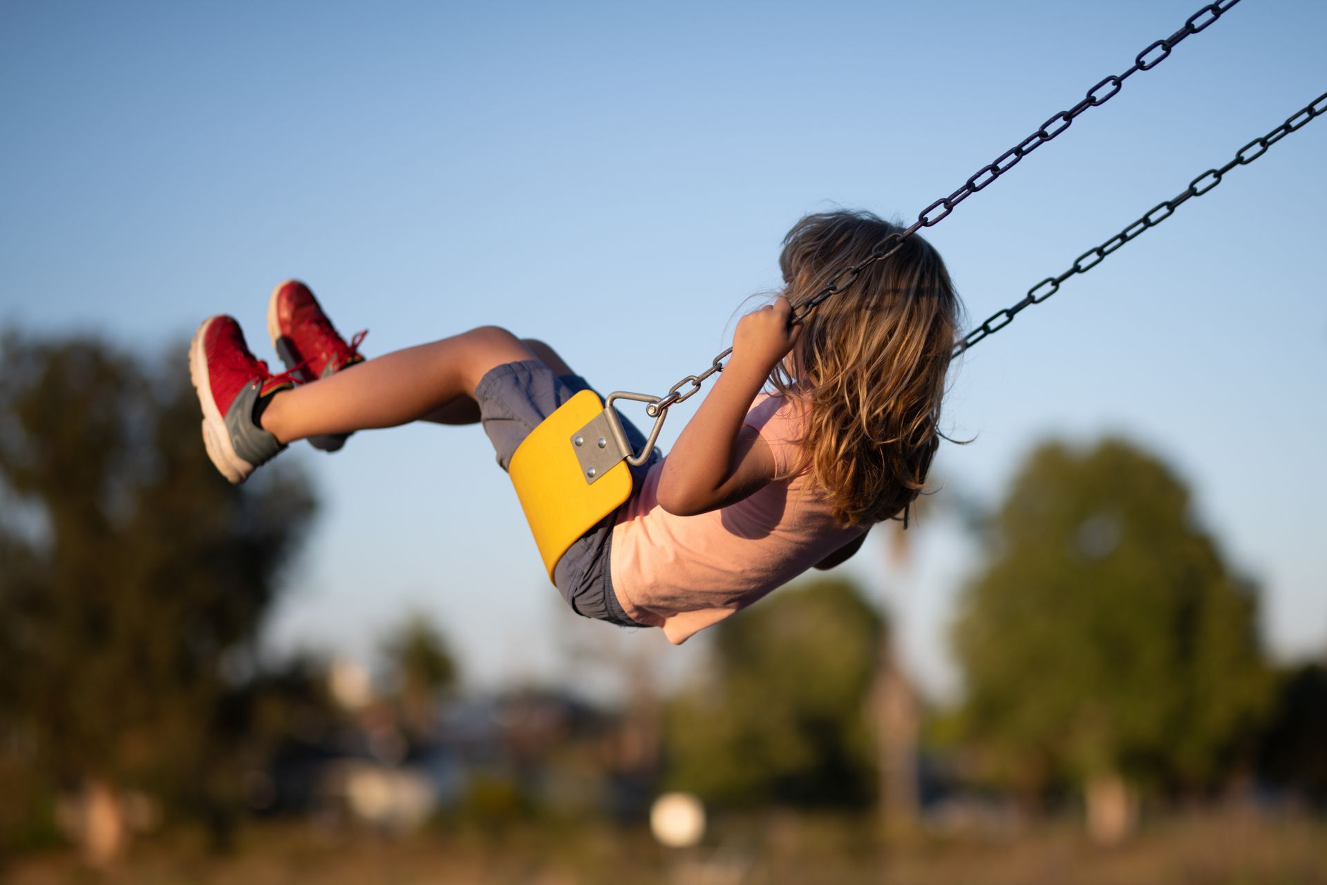 Child swinging high in the air, wearing red shoes. Set against a blurred, sunny outdoor background.