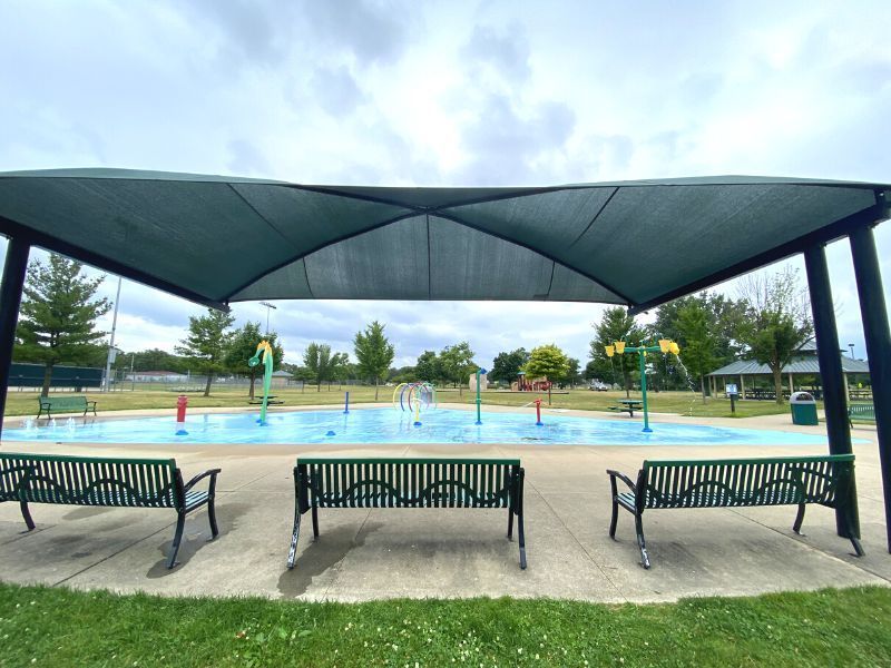 A splash pad with water features, benches, and a shade canopy under a cloudy sky.