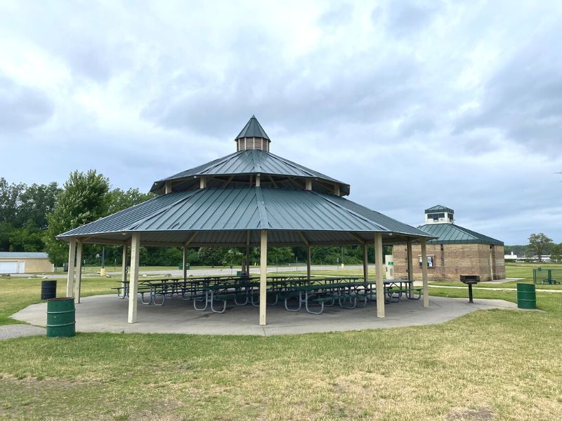 A green-roofed gazebo with picnic tables, on a grassy area under a cloudy sky.