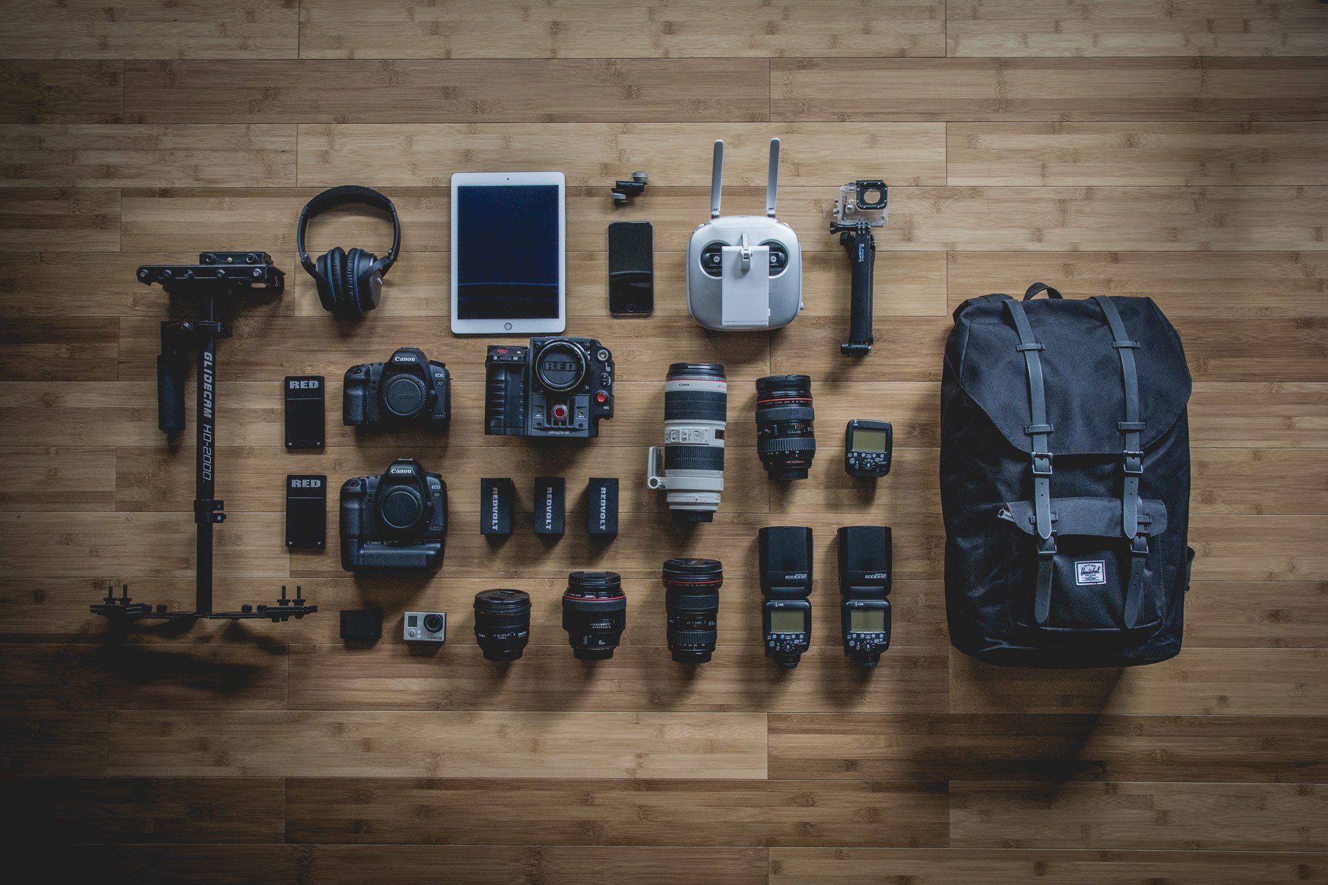 A bunch of cameras , lenses , headphones , and a backpack are sitting on a wooden floor.