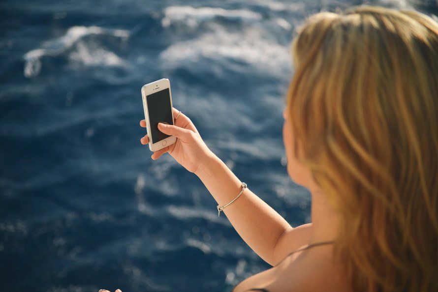 A woman is taking a selfie with her cell phone in front of the ocean.