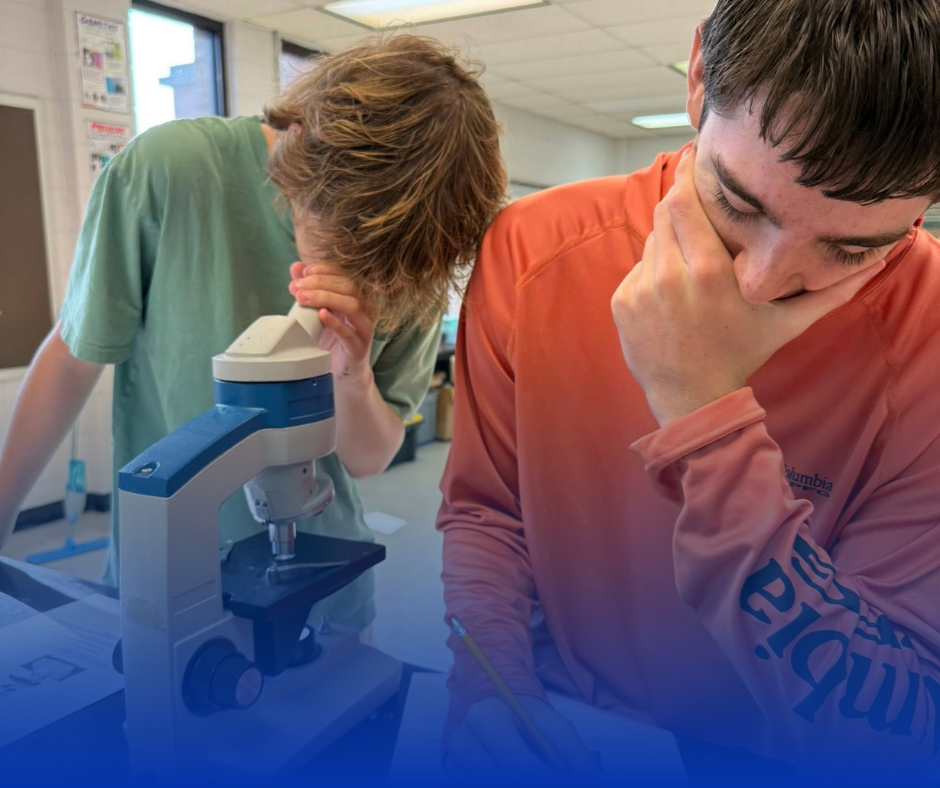 Two students in a lab; one looks through a microscope, the other ponders with hand on his face.