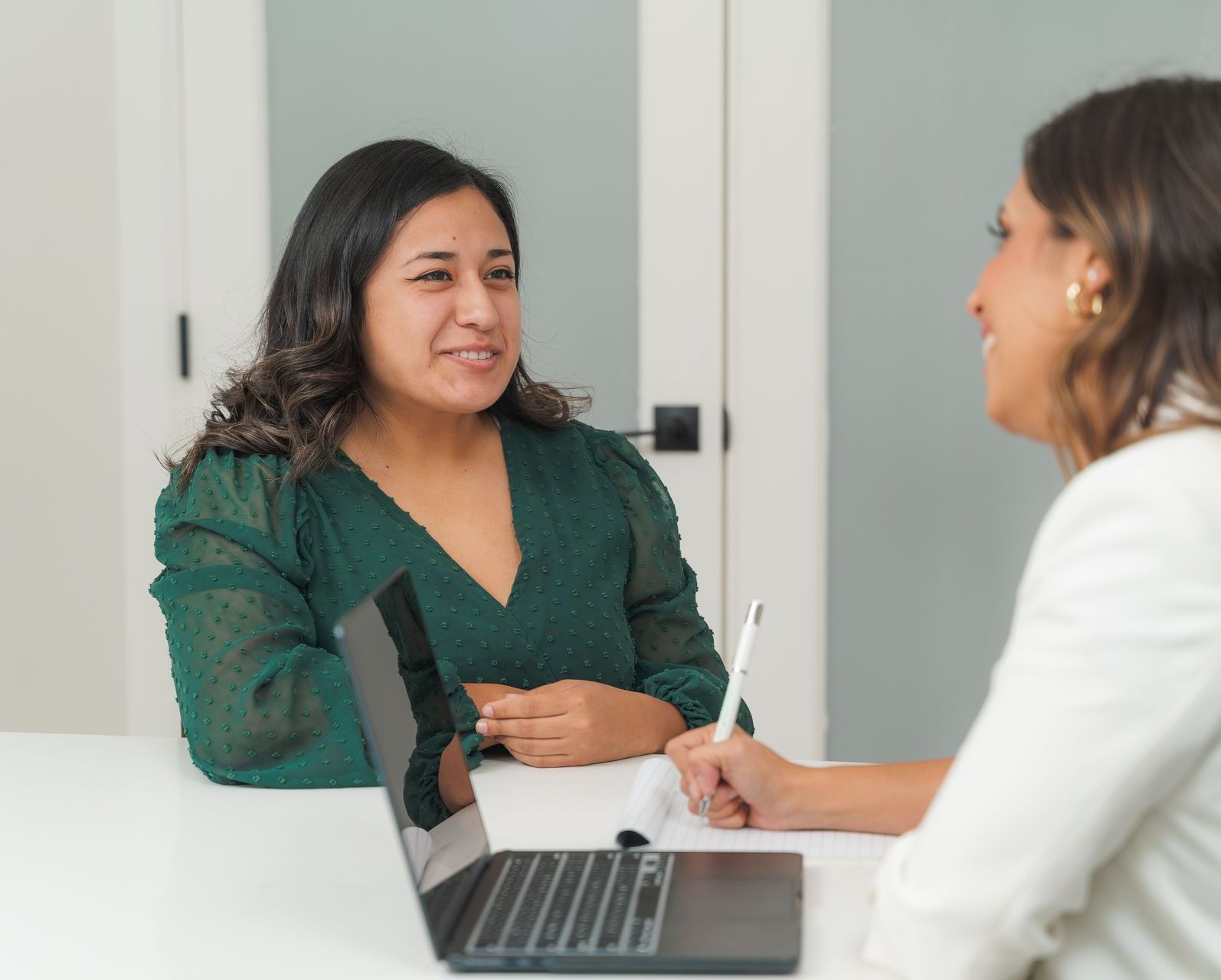 Two women are sitting at a table with a laptop and talking to each other.