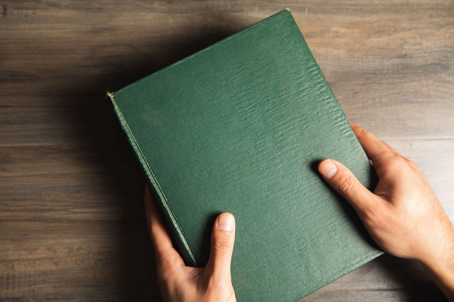 A person is holding a green book in their hands on a wooden table.