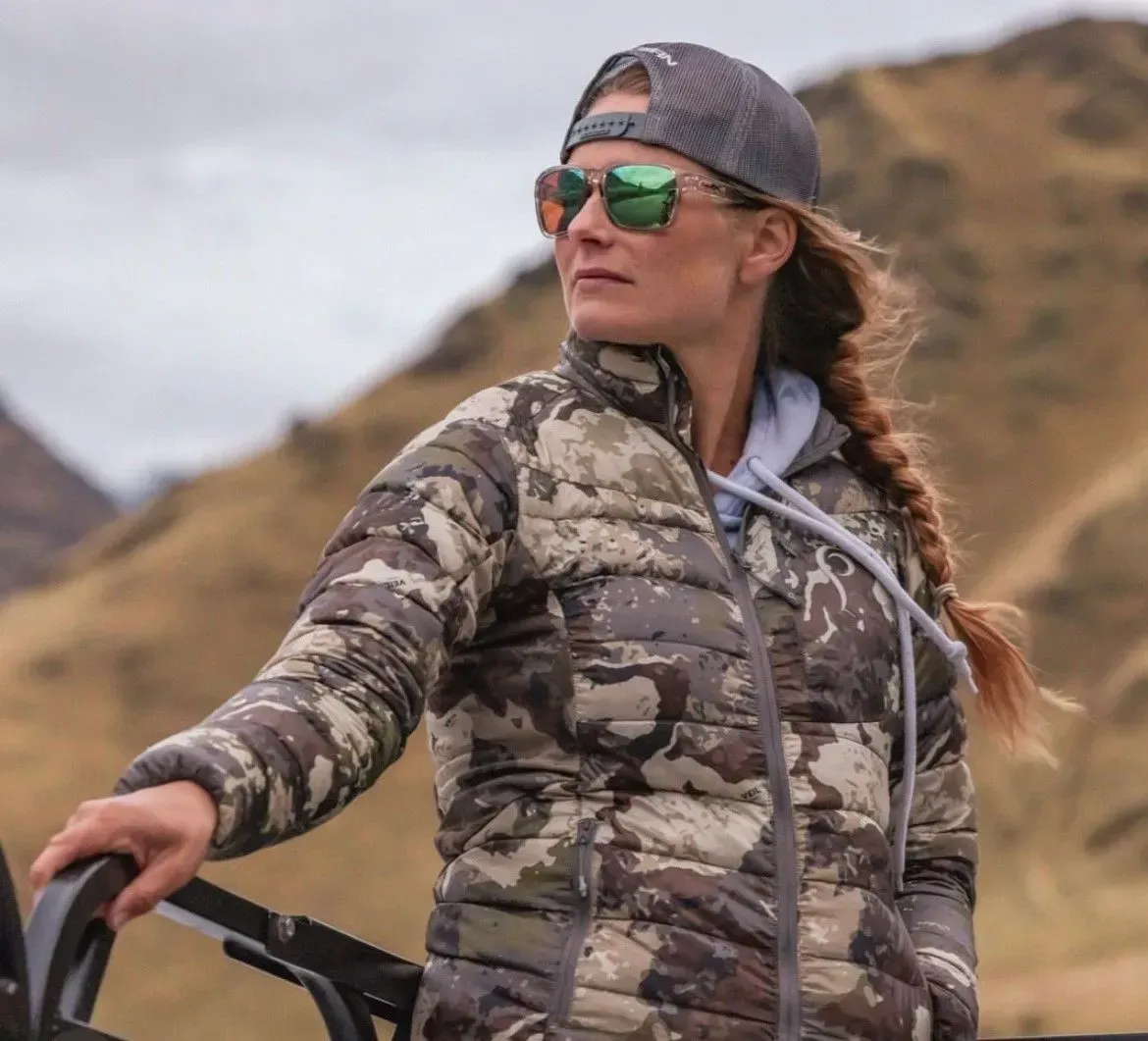Woman in camo jacket, sunglasses, and baseball cap looking off into the distance, near a hilly landscape. — EyeMax EyeCare in Singleton, NSW