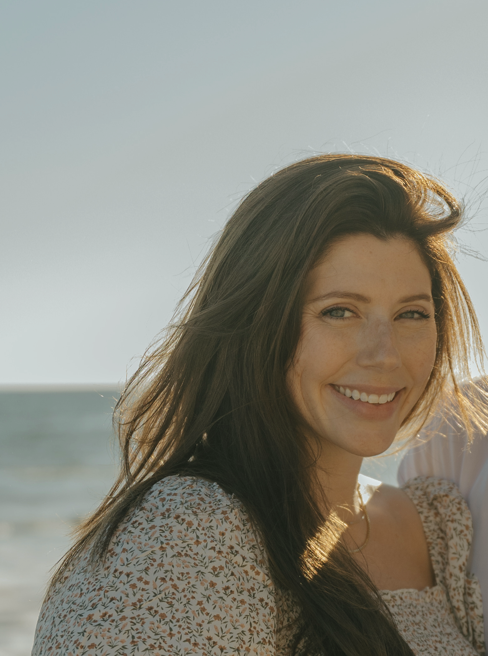 Woman with long brown hair smiling on the beach, wearing a light-colored patterned sweater, with the ocean in the background.