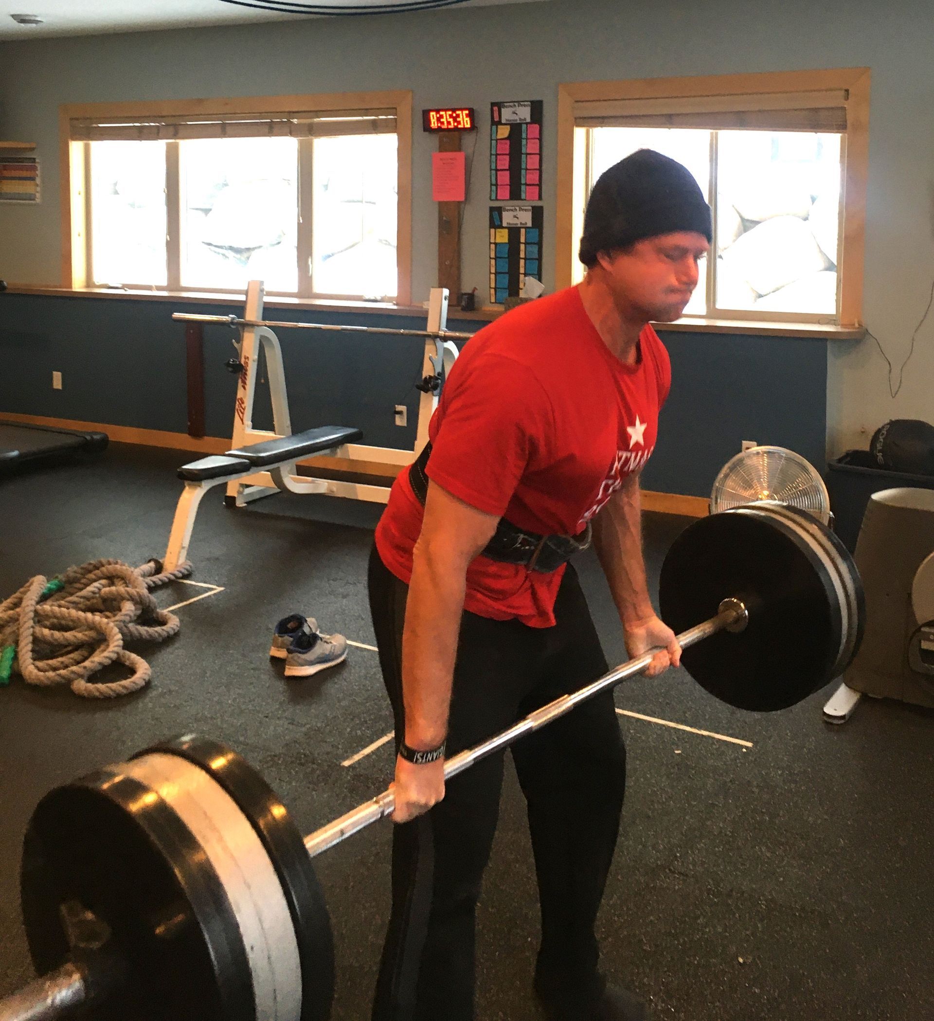 Man in red shirt deadlifting a barbell in a gym, wearing a black beanie and weightlifting belt.