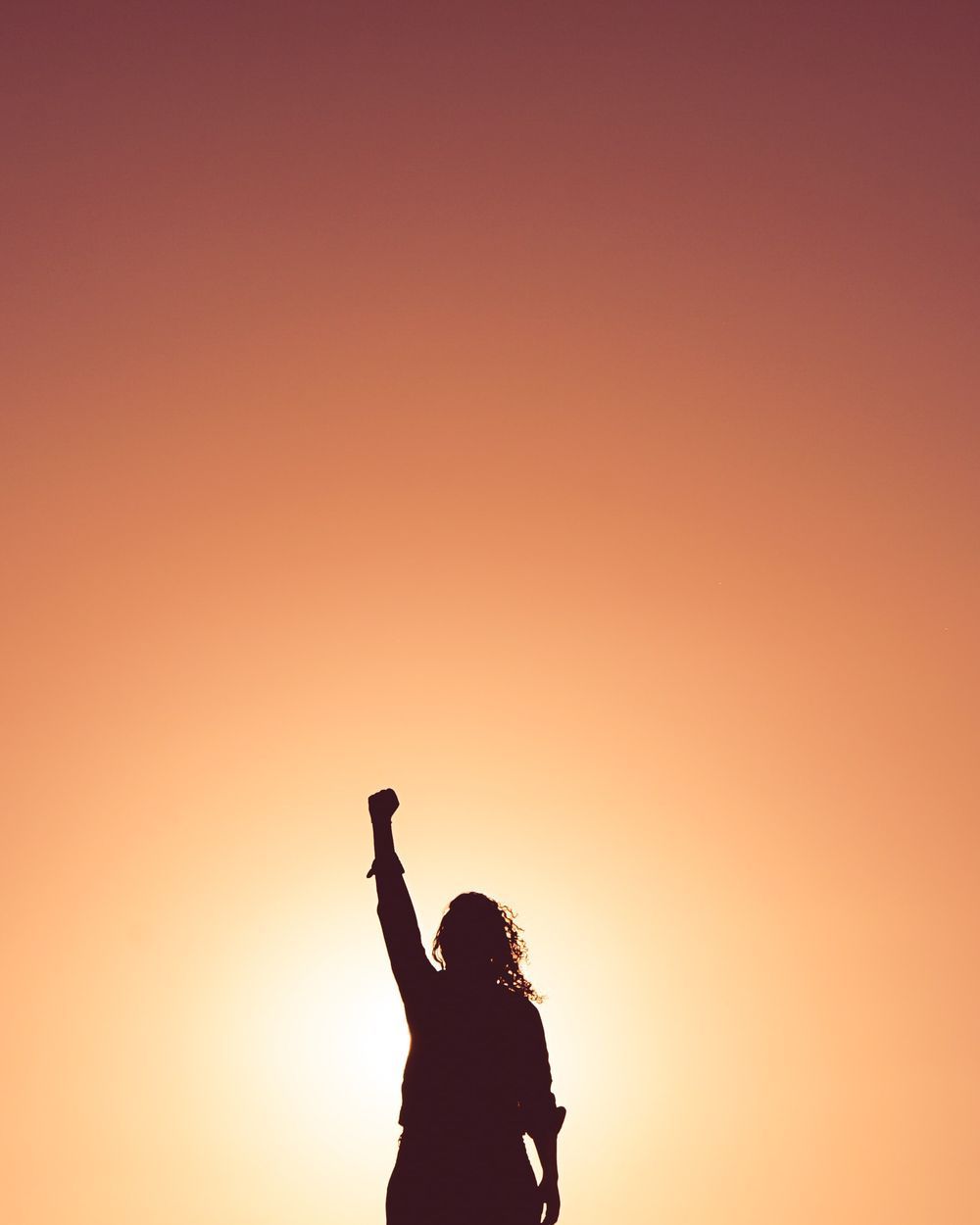 A silhouette of a woman raising her fist in the air at sunset.