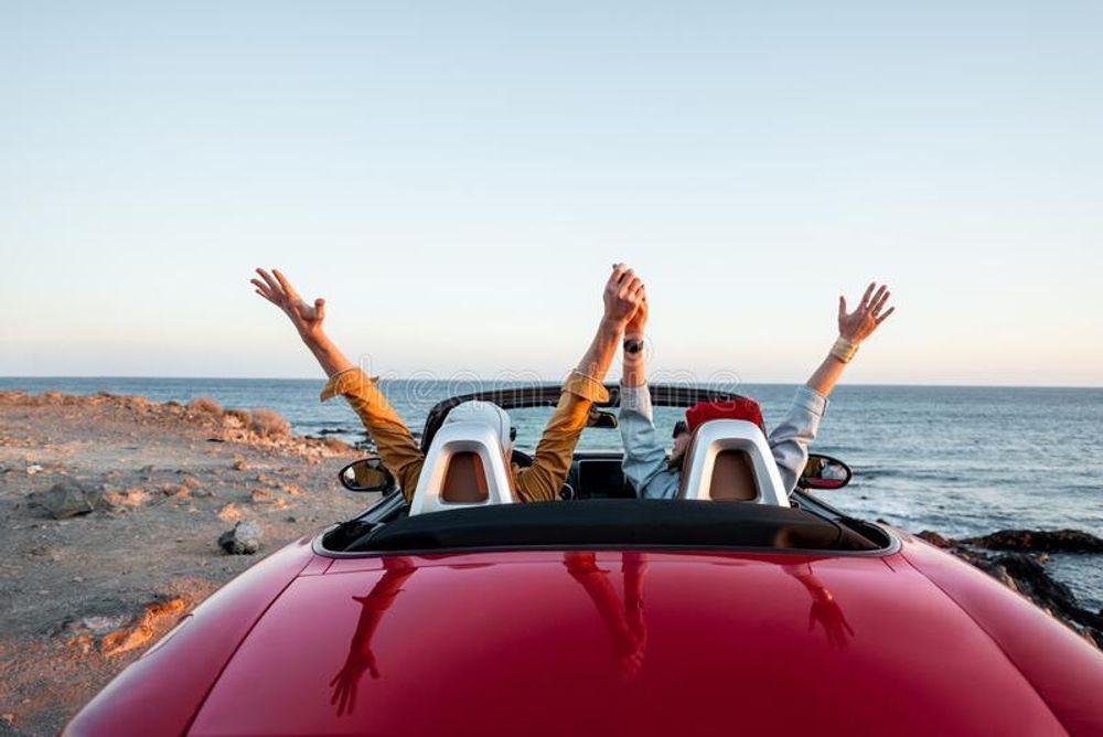 Two people are sitting in a red convertible car with their arms in the air.