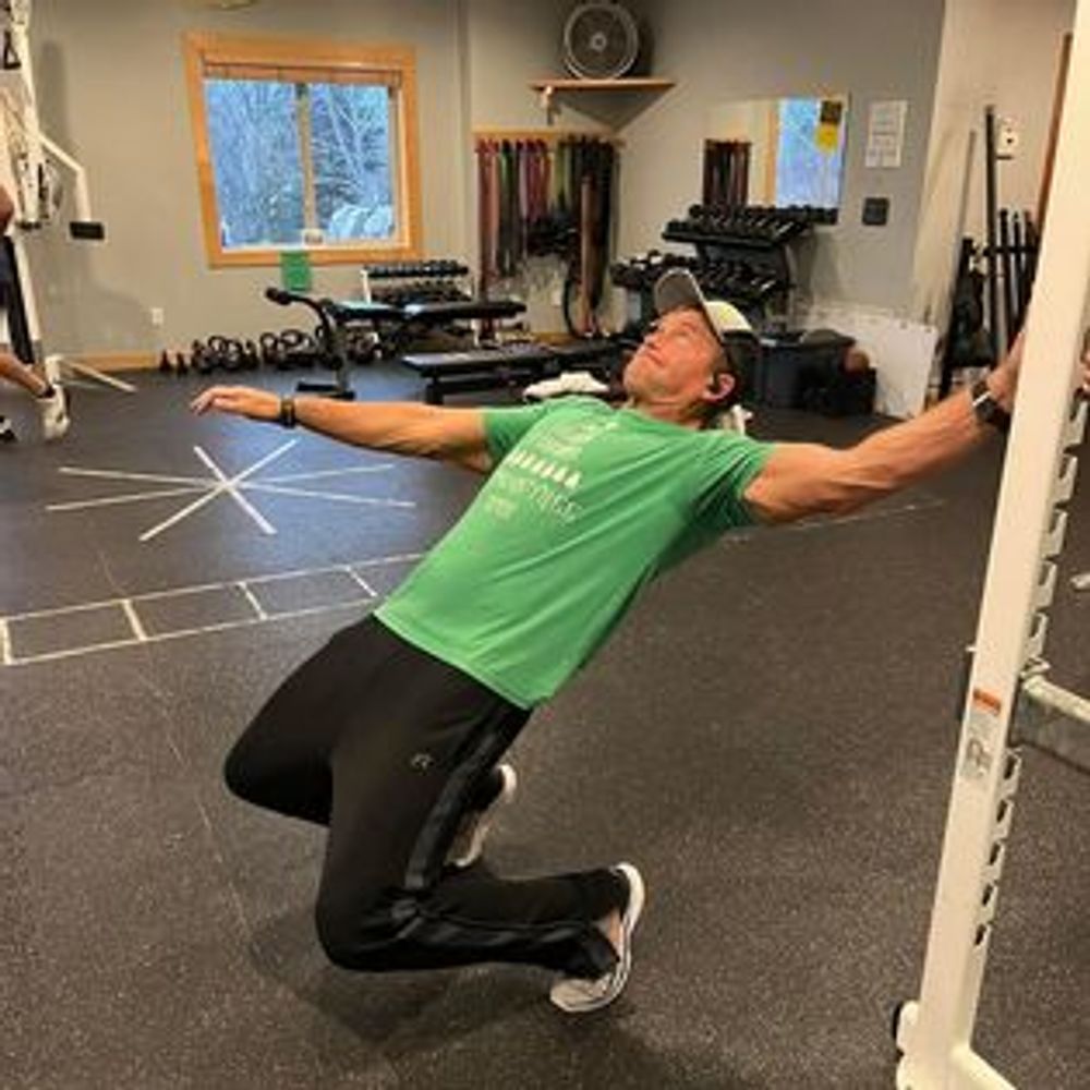 A man in a green shirt is kneeling down in a gym with his arms outstretched.