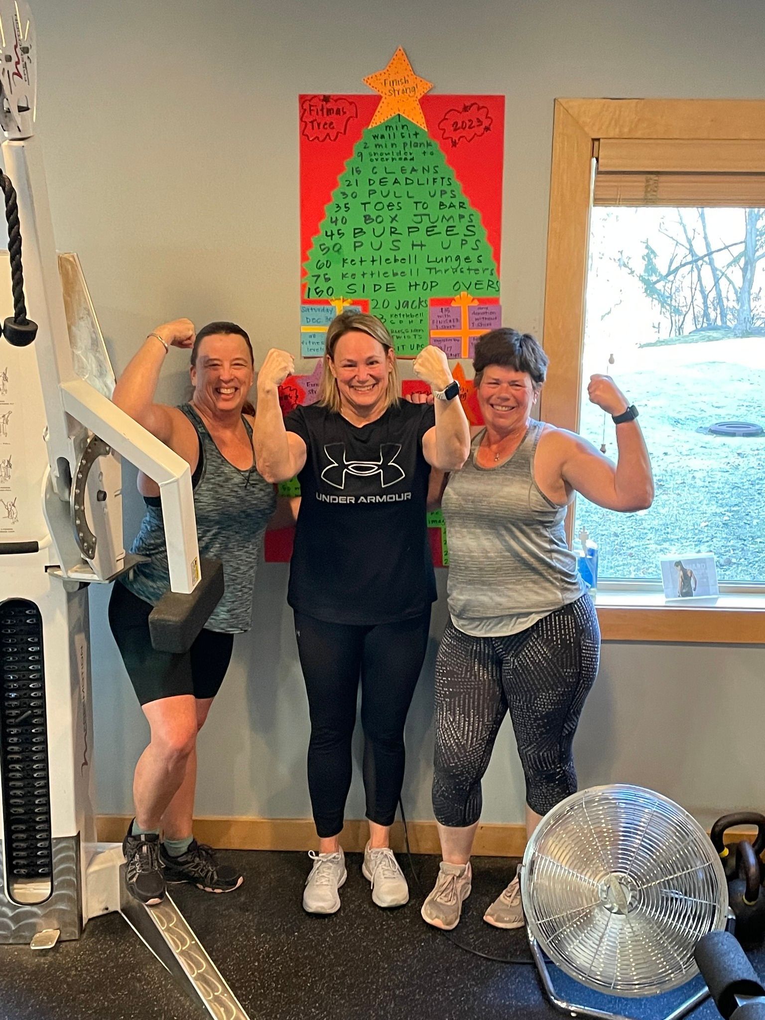 Three women are posing for a picture in a gym in front of a christmas tree.