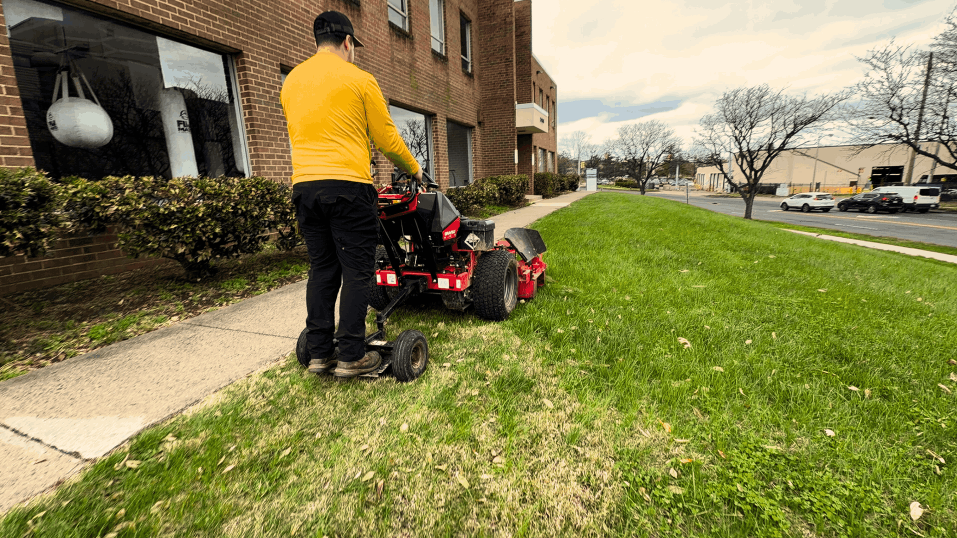 A person in a yellow sweater stands on a red stand-on lawn mower, cutting grass beside a brick building.