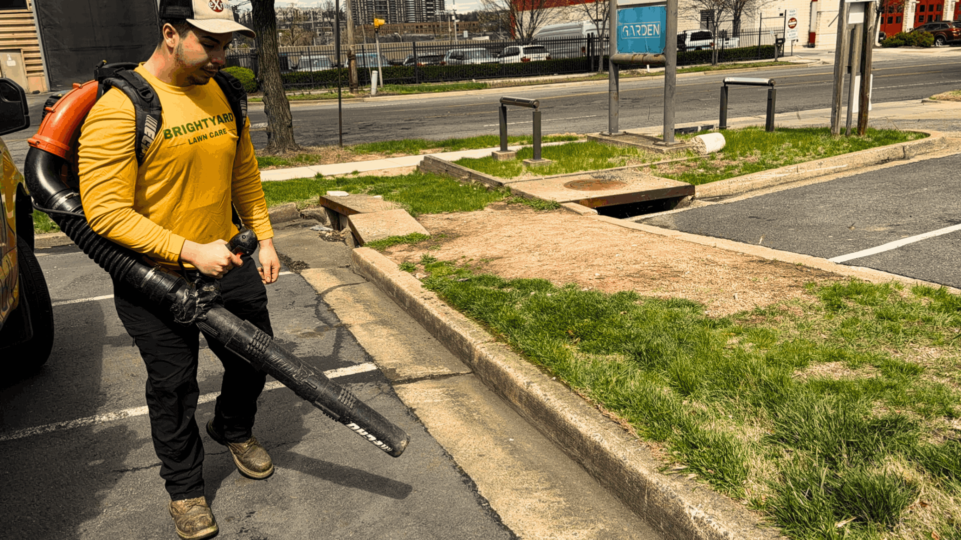 A worker in a yellow long-sleeved shirt uses a backpack leaf blower on a paved parking lot near a grass verge.