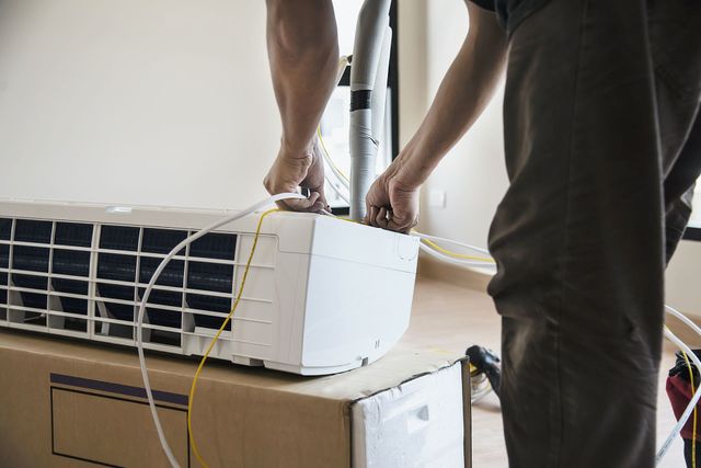Person working on a white window air conditioner with tools and wires nearby indoors