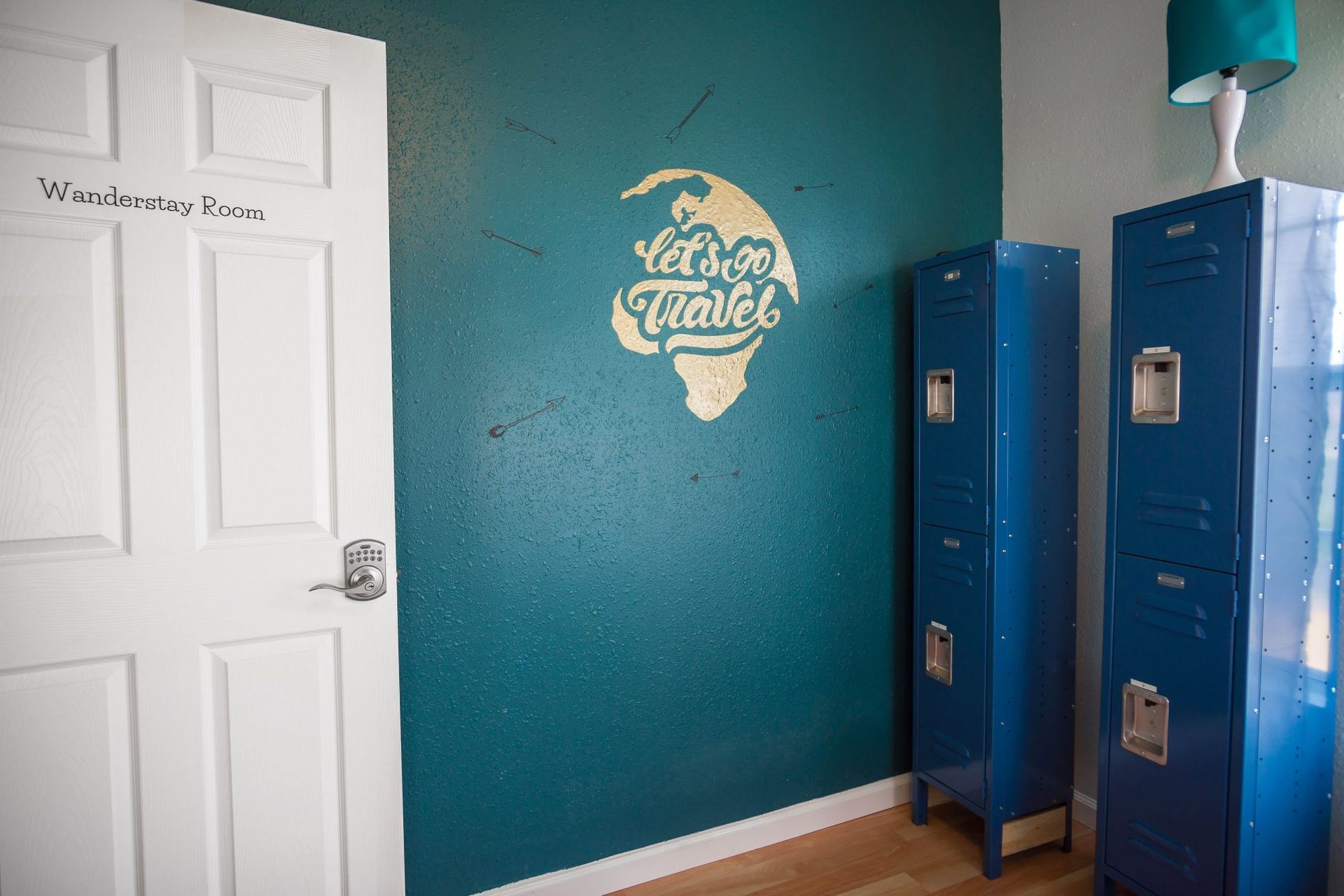 A teal wall with travel-themed art and two blue lockers. A white door is on the left.