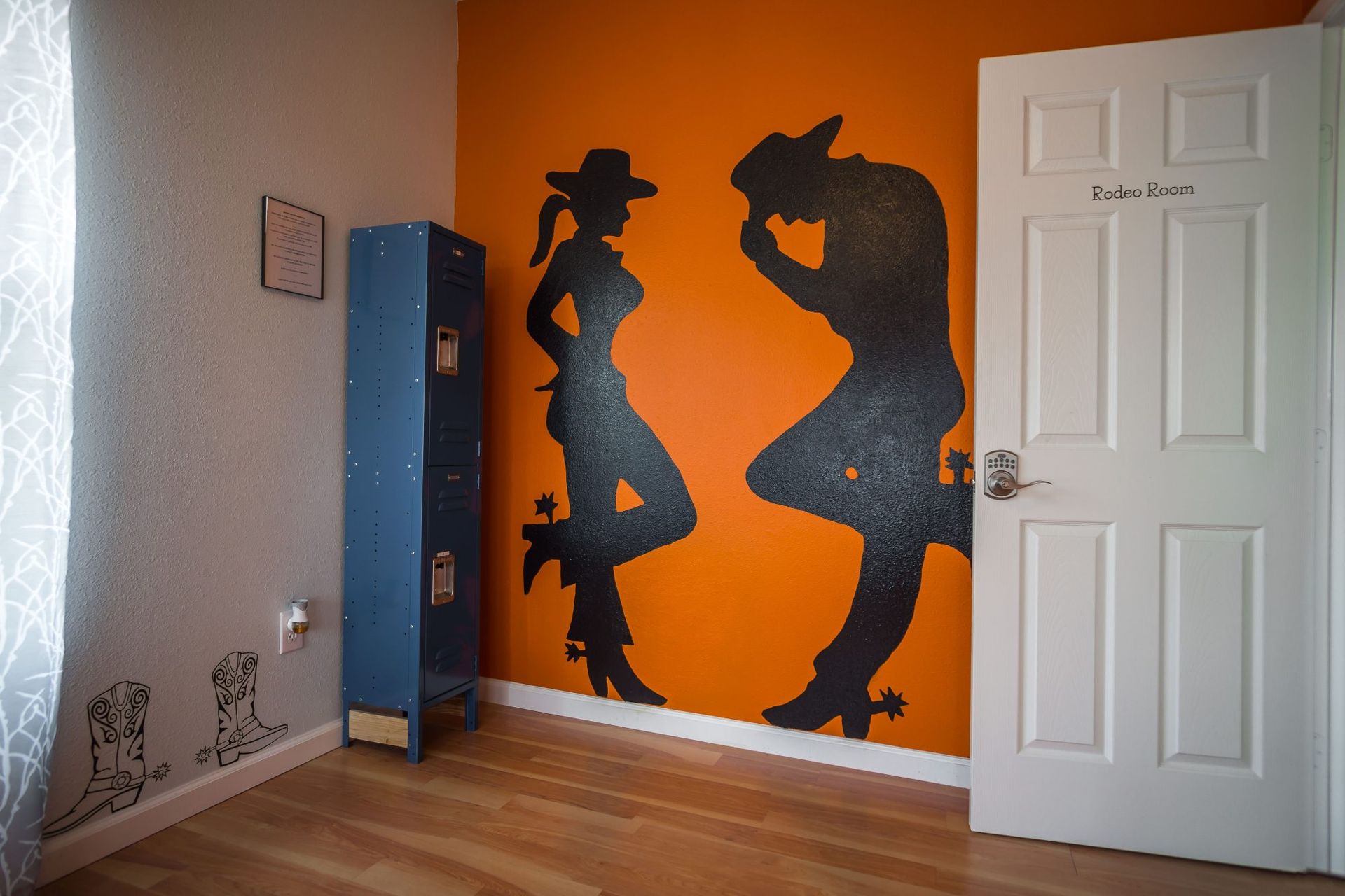 A room with orange accent wall featuring a cowboy and cowgirl silhouette, blue locker, and white door.