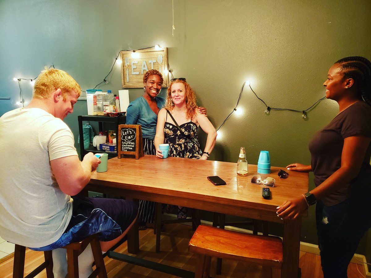 Four people around a wooden table. One sits, three stand, all smiling, fairy lights on wall.