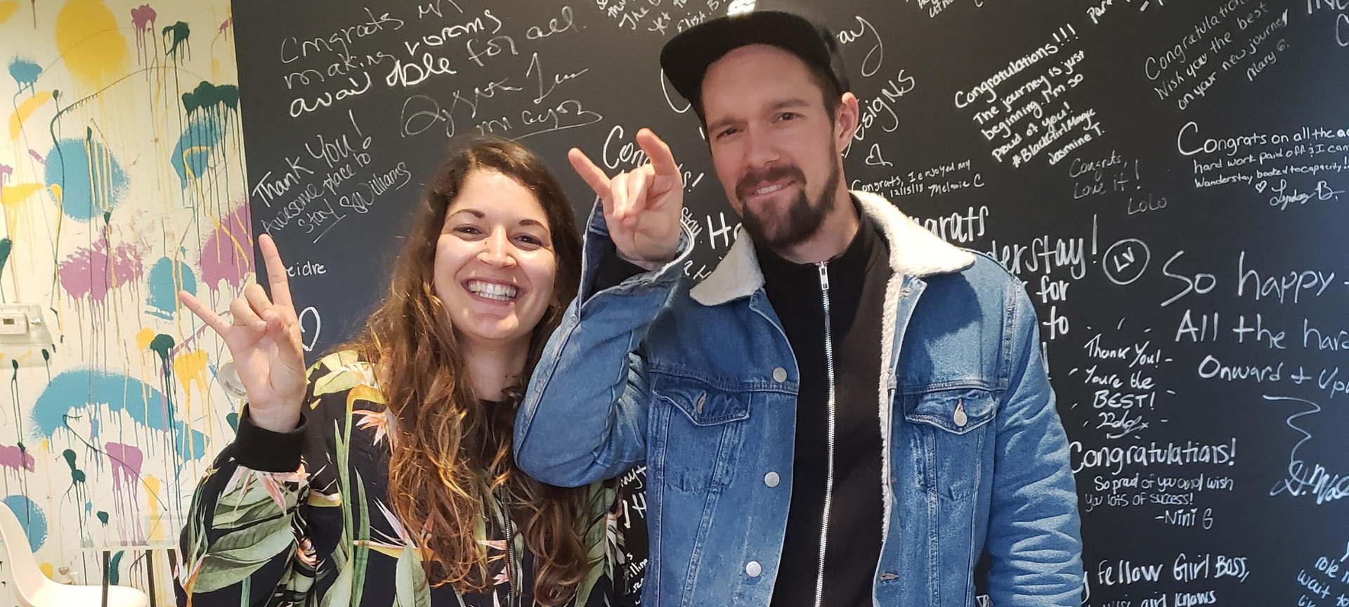 Two people, smiling, giving peace signs. They stand near a wall with writing.