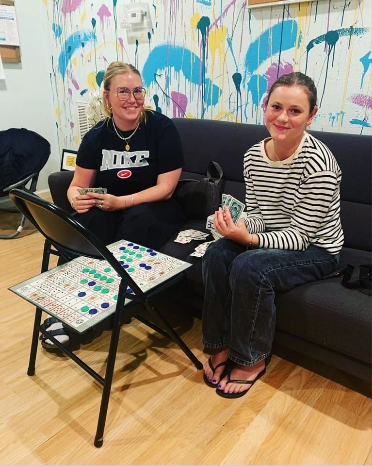 Two people playing a game. One sits by a chair with tiles, the other on a couch holding cards. Colorful wall background.