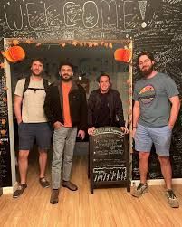 Four men stand in a doorway with pumpkins, welcome sign above. Two men hold a chalkboard.