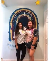 Two people stand in front of a painted arch that says, 