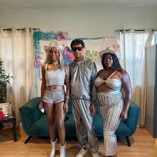 Three people in futuristic silver outfits pose in front of a blue couch and decorative backdrop.