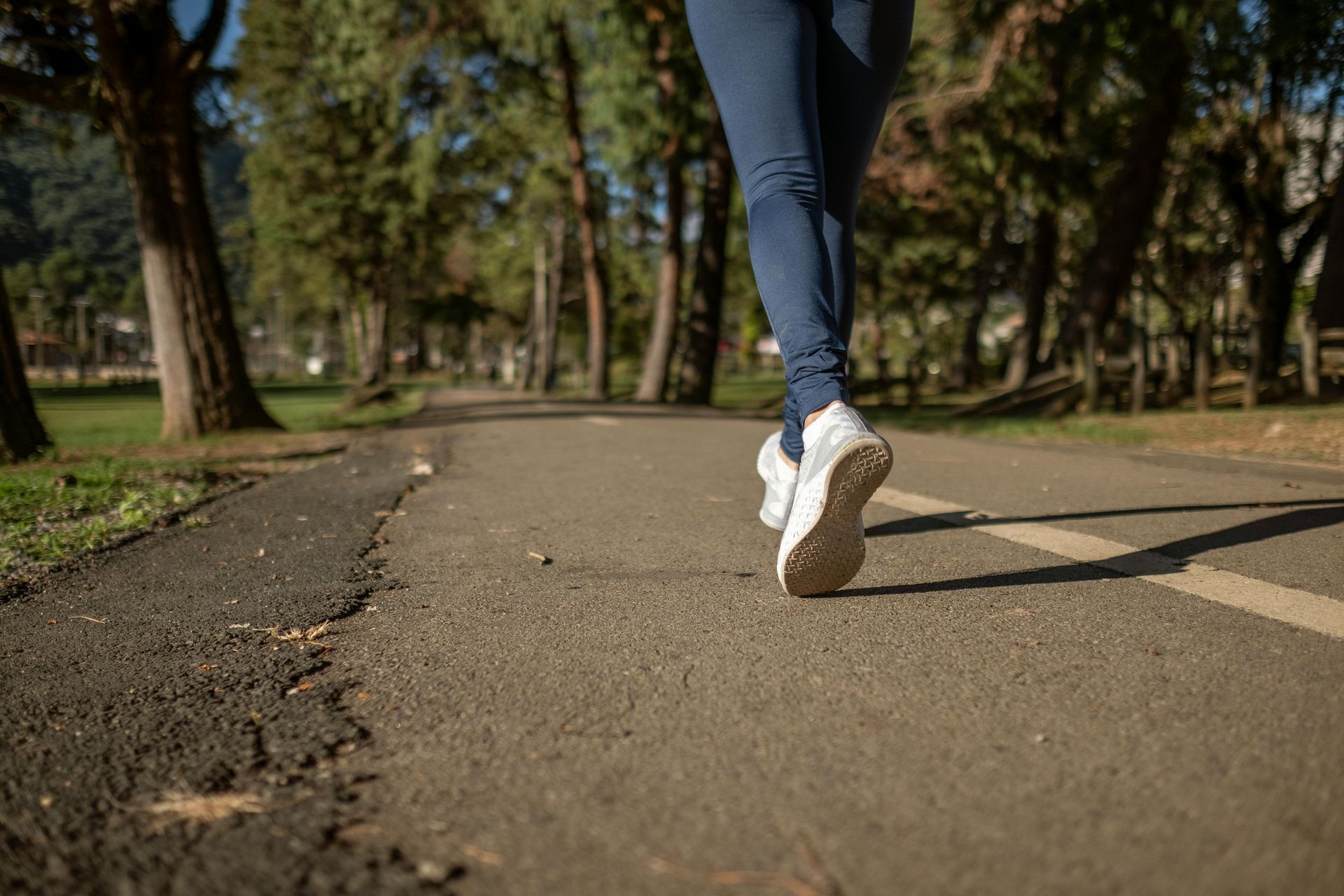 Runner in blue leggings and white shoes on a paved path in a park with trees.