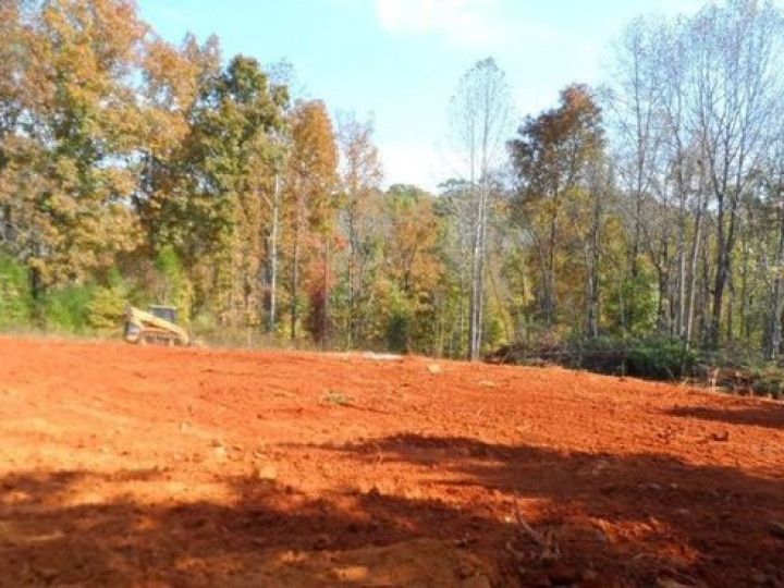 Red dirt clearing with trees in the background and a yellow construction vehicle on the left.
