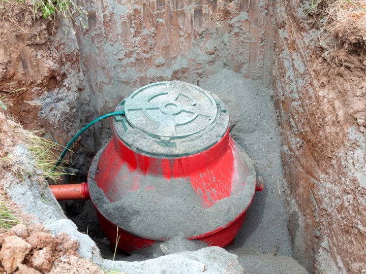 Red septic tank installed in a dirt pit, surrounded by sand, with lid and connected pipe.