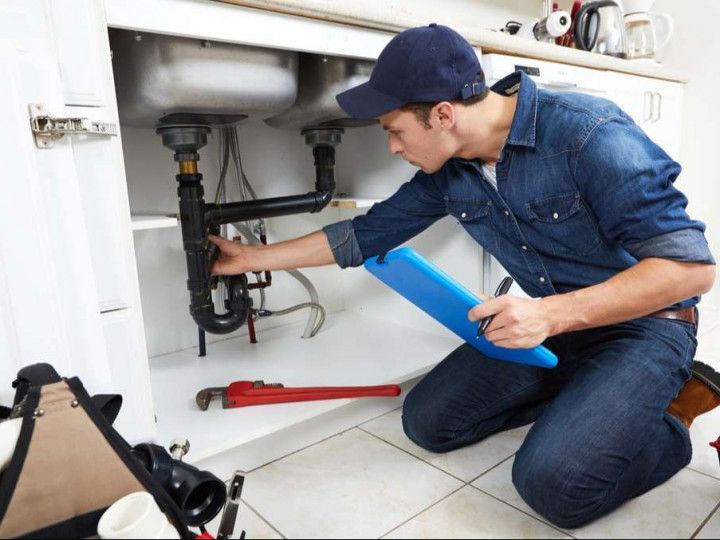 Plumber kneeling under a kitchen sink, inspecting pipes. He wears a blue hat and holds a clipboard.