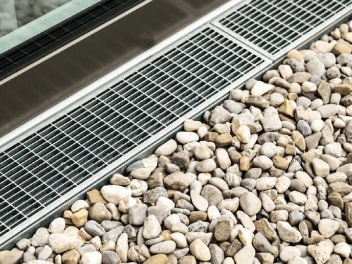 Metal grate drain along a building, next to a bed of pebbles.