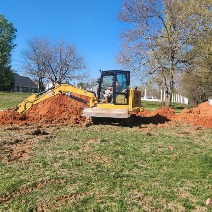 Yellow excavator digging in a grassy field with mounds of red soil, under a blue sky.