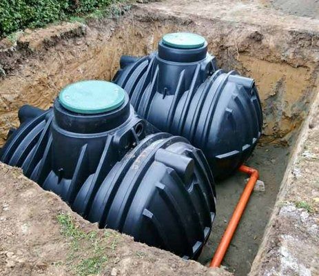 Three black septic tanks in an excavated pit, connected by orange pipes, in a yard.