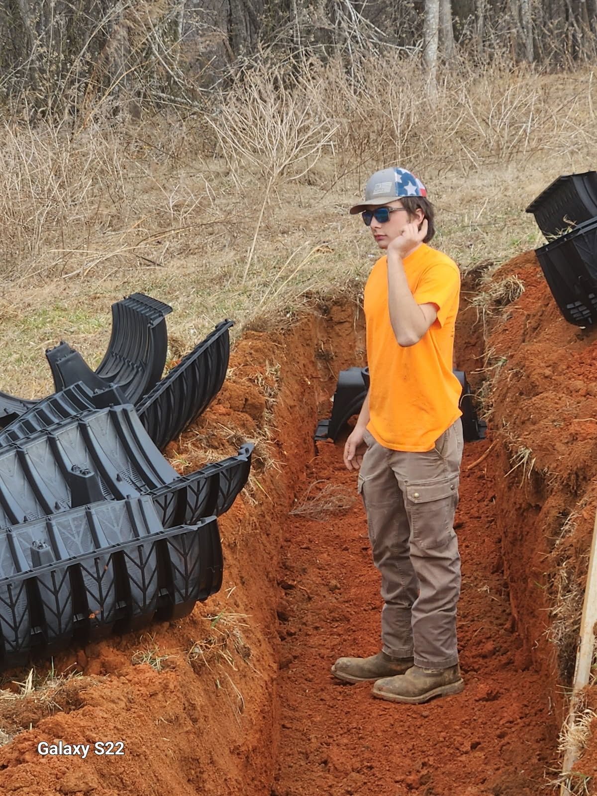Person in orange shirt and hat in a trench, looking at a septic tank component in a field.