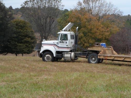 White semi-truck with trailer parked in a grassy field, trees in the background.