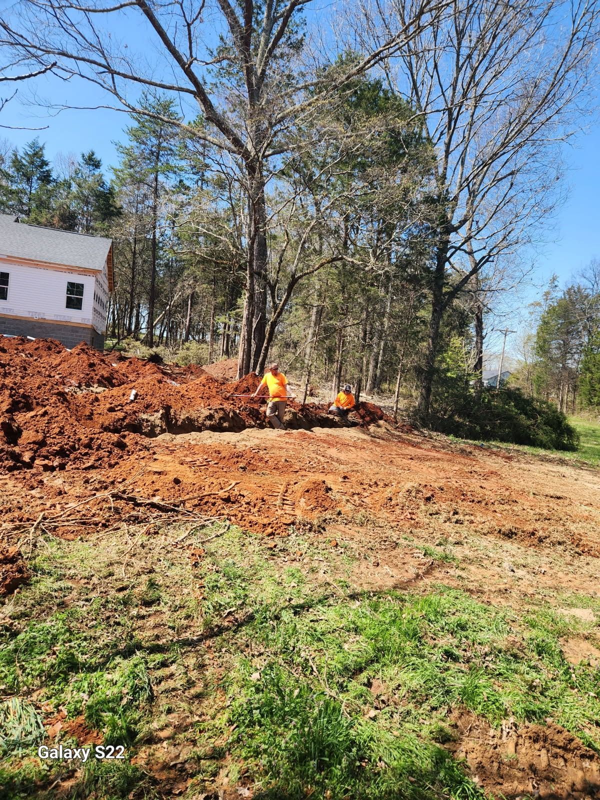 Construction site with two workers in orange vests, red soil, trees, and a house under construction.