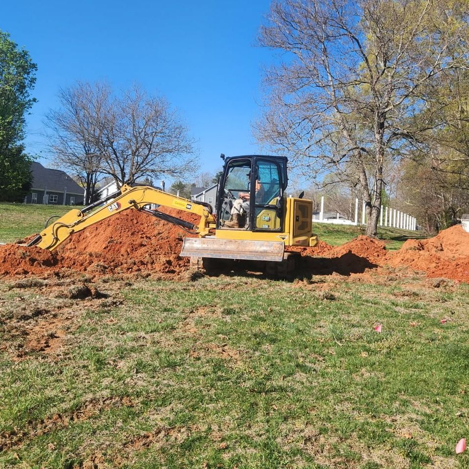 Yellow excavator digging on a grassy field; dirt piles nearby under a blue sky.