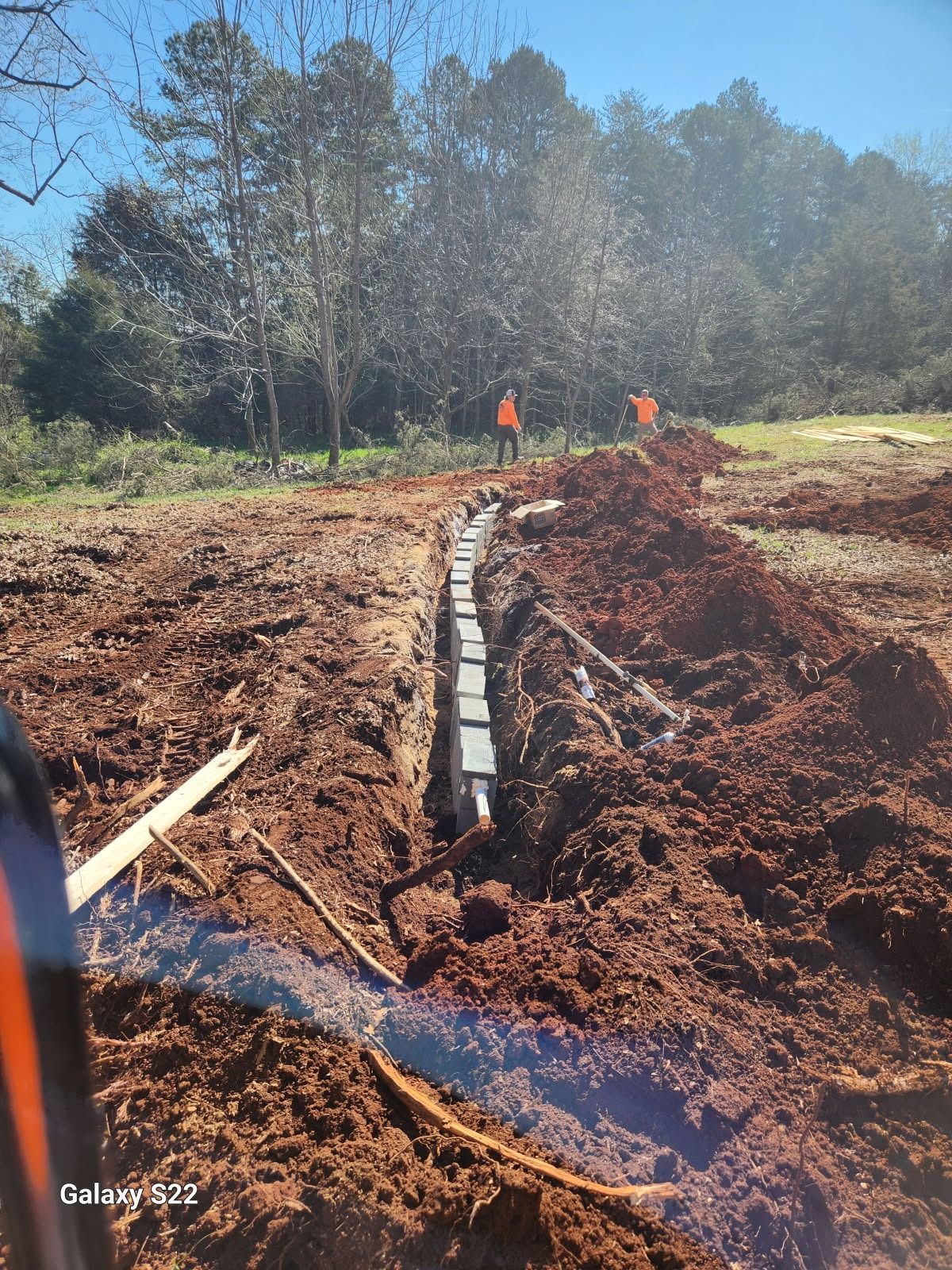 Ditch with buried pipes, workers in orange vests; dirt, trees in background, sunny day.