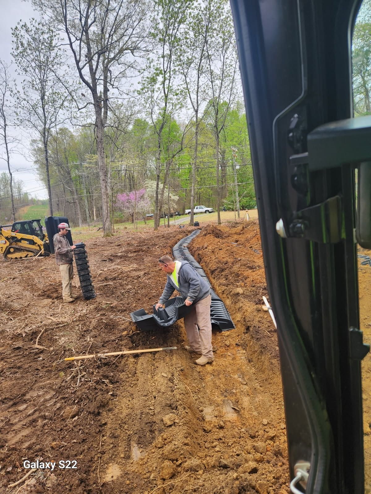Two workers installing black drainage pipe in a ditch at a construction site. Trees in the background.