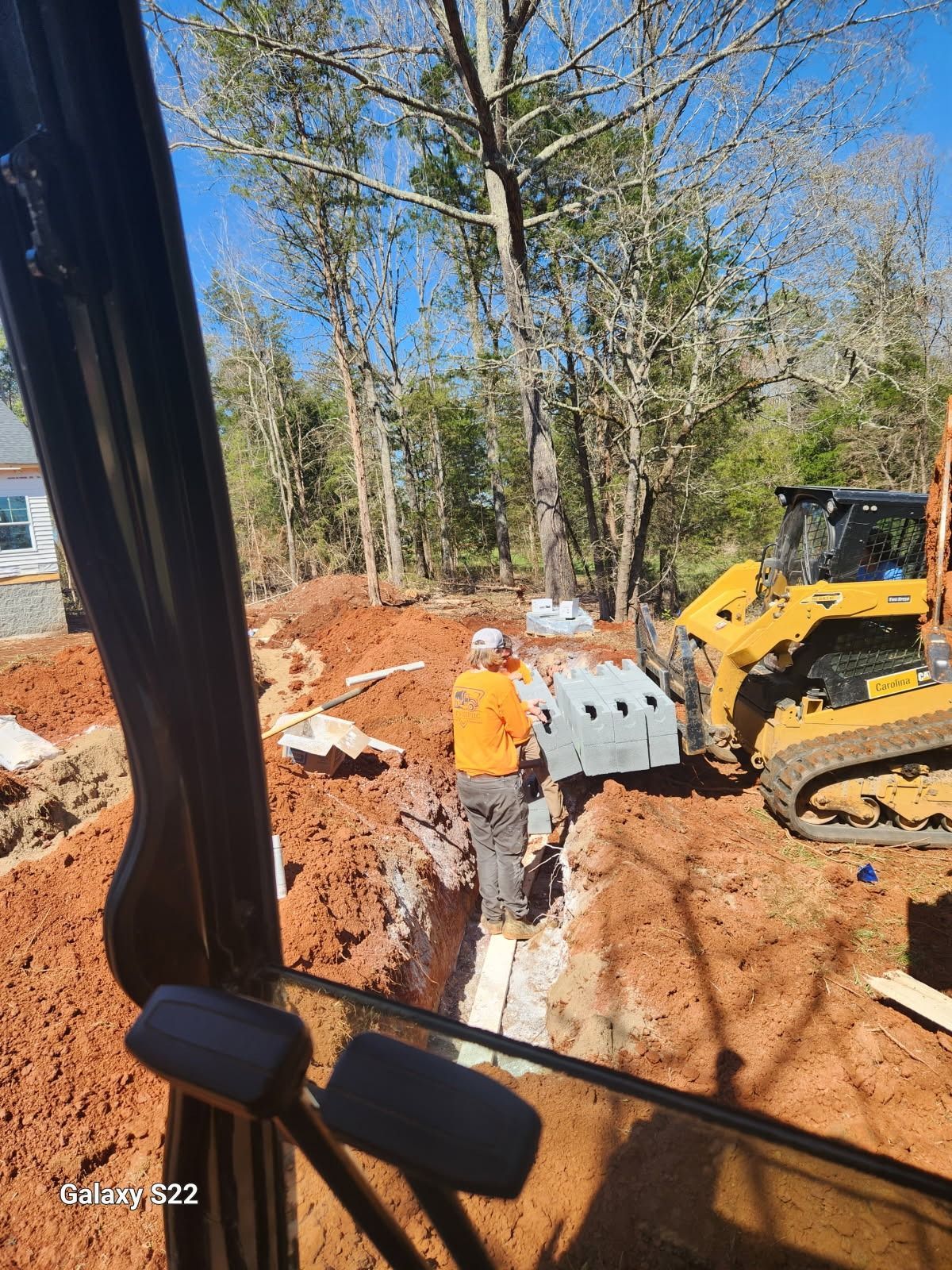 A construction worker in an orange shirt standing in a trench. A yellow skid steer and construction materials nearby.