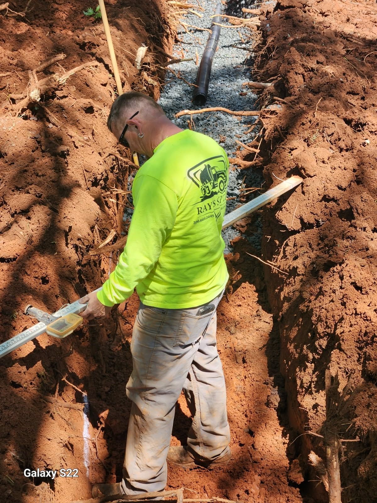 Construction worker in neon green shirt checks pipe level in a trench.