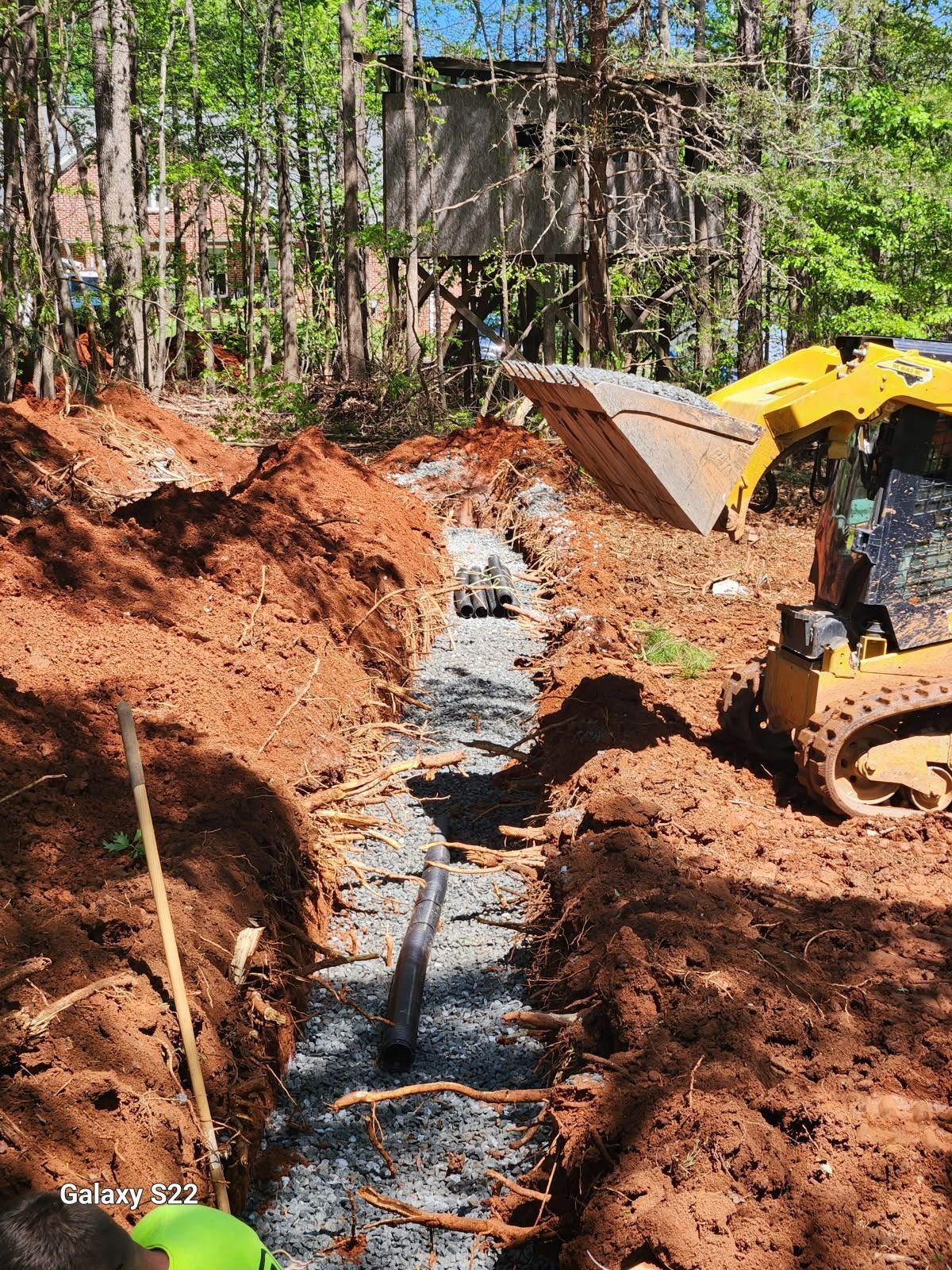 A trench filled with gravel and pipe, alongside a small excavator, near a wooded area.